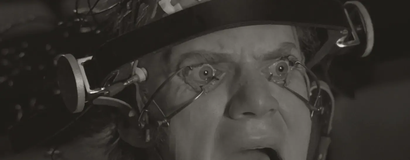 Film still: a man’s eyes held open by clamps